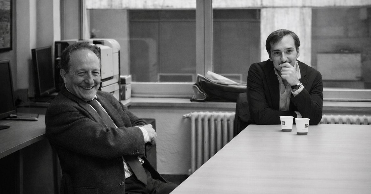 Fotografía en blanco y negro: Rafael Alvira, a la izquierda, ríe sentado junto a una mesa; a la derecha, Alberto Ortiz lo escucha con la mano en la barbilla. Sobre la mesa hay dos vasos de café.